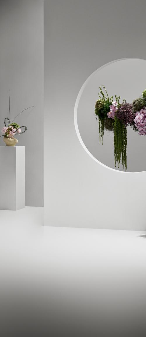 A smartly dressed woman of color walking across a room scattered with intricate floral arrangements. The arrangements are placed around various architectural structures of the space - an archway, a circular window, a doorway.